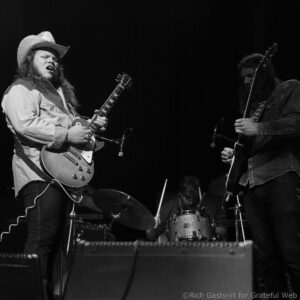 Two guitarists perform on a black-and-white stage; left musician wears a cowboy hat and plays a sunburst guitar, right musician focuses on his guitar behind a drum set.