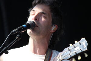 Close-up of a male singer with eyes closed, singing into a microphone with a guitar neck in the foreground.