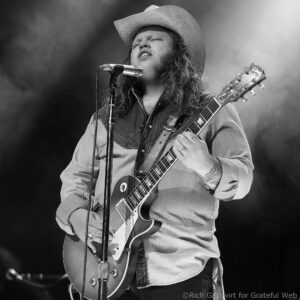 Guitarist in a denim shirt and wide-brim hat sings into a microphone while playing an electric guitar on stage.