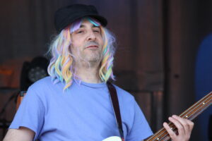 Man with pastel rainbow wig and black cap playing a bass guitar on stage, wearing a light blue T-shirt