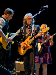 Three musicians perform on a dark stage, each with an electric guitar: left guitarist in a striped shirt and vest, center guitarist with long gray hair and a yellow guitar, right guitarist in a floral dress.