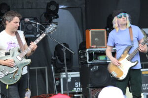 Two guitarists perform on a stage: left, a man in a white tank top playing a pale-green electric guitar; right, a man with a pastel wig and blue shirt playing a bass guitar with sunglasses.