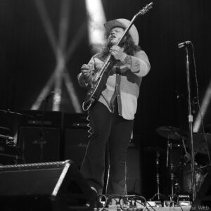Male guitarist with long hair and a cowboy hat playing an electric guitar on stage under a spotlight.