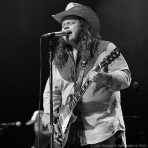 Male guitarist wearing a straw cowboy hat and denim jacket sings into a microphone while playing an electric guitar on stage.