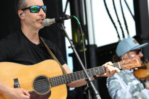 Male guitarist singing into a microphone while playing an acoustic guitar outdoors, wearing sunglasses and a black shirt, with another musician in a camo hoodie in the background