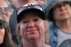Smiling woman in a black cap with sunglasses on her head, close-up outdoors, other people blurred in background (group photo)