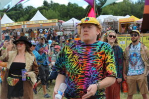 Man in a bright tie-dye shirt and yellow cap at an outdoor festival, holding a water bottle, with vendor tents in the background.