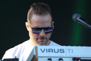 Male musician wearing reflective sunglasses, close-up on his face, performing on stage with a microphone to the right and a sign in the foreground.