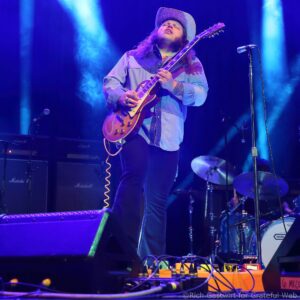 Male guitarist wearing a cowboy hat and denim shirt, playing a sunburst electric guitar on a blue-lit stage.