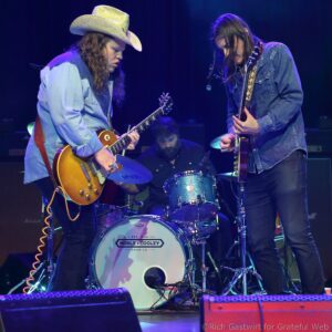 Two guitarists in denim jackets perform on a dimly lit stage, one wearing a cowboy hat; a drummer sits behind them.