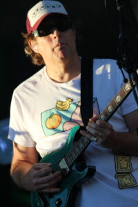 Guitarist wearing a white and red cap and sunglasses playing a teal-green electric guitar on stage.
