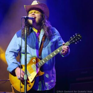 Male singer wearing a straw cowboy hat and denim shirt, singing into a microphone while playing a sunburst Les Paul guitar on stage under colorful lighting.