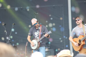 Two guitarists perform on a stage; the left musician wears a pink hat and sunglasses while playing a banjo, the right musician plays an acoustic guitar with sunglasses visible in the foreground and audience out of focus.