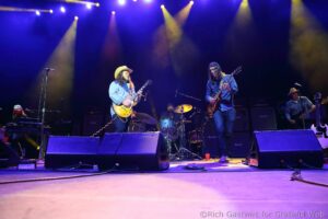 Live band on stage playing guitars under blue and yellow spotlight beams; drummer in back, keyboardist left, cowboy-hatted guitarist center-left in denim shirt, another guitarist right.