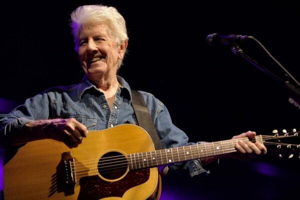 Older man in a denim shirt playing an acoustic guitar on a dark stage, smiling as he performs.