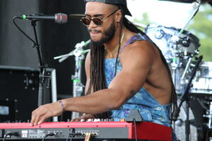 Musician with dreadlocks wearing sunglasses and a backward cap, playing a red keyboard on stage with a microphone nearby.
