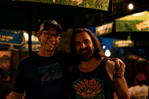 Two men posing with arms around each other at a nighttime outdoor venue, smiling; one wears a cap and glasses, the other has long hair and a tattooed arm in a sleeveless shirt.