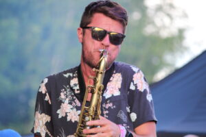 Man wearing sunglasses plays a saxophone outdoors with a floral shirt, blurred greenery in the background.
