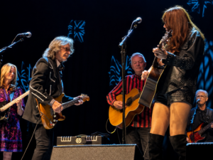 A rock band performs on stage: guitarist in black with a sunburst strat, vocalist/guitarist in studded leather, and a striped-shirt guitarist in the background.