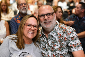 Smiling couple close-up at a social event, woman in gray shirt and man in a floral shirt, with a blurred crowd in the background.