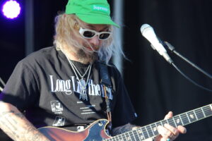 Guitarist wearing a bright green cap and white sunglasses plays an electric guitar on stage, microphone nearby, purple stage lights in the background.