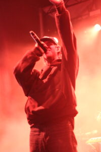 Singer on stage wearing a cap, mic in hand, raised fist under red stage lights.