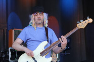 Musician with rainbow wig and black cap playing a white Fender bass on stage, eyes closed under blue lighting.