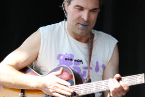 Close-up of a man in a sleeveless shirt playing an acoustic guitar, fingers on the fretboard.