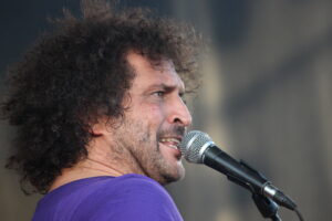 Close-up of a man with curly hair singing into a microphone, wearing a purple shirt.