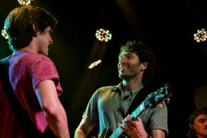Two male musicians on stage share a smile while one plays an acoustic guitar, colorful lights overhead.