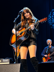 Woman on stage playing a sunburst acoustic guitar and singing into a microphone, with a bassist in the background.
