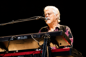 Older man with white hair and glasses singing into a microphone while playing a keyboard on stage, wearing a dark patterned shirt.