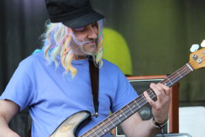 Musician with rainbow wig and black cap playing a bass guitar in a blue shirt on stage.