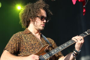 Musician with curly hair wearing a patterned shirt playing an electric guitar on stage under green lights