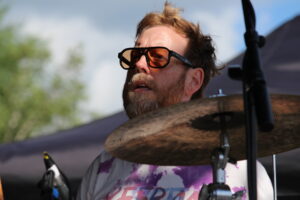 Bearded man wearing orange-tinted sunglasses playing drums on stage, partially visible cymbal in foreground.
