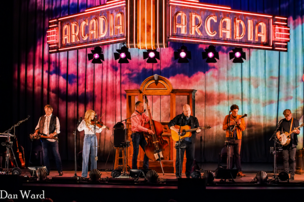 Six musicians perform on a colorful stage beneath a bright Arcadia Arcade neon sign.
