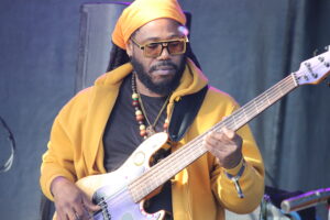 Musician in an orange head wrap and yellow hoodie playing a bass guitar on stage.