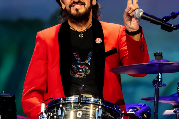 Drummer on stage in a red blazer and sunglasses, flashing a peace sign while playing a silver drum kit.