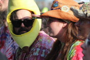 Two festival-goers wearing tie-dye shirts; one in a yellow helmet and sunglasses, the other in a straw hat with pins and face paint.