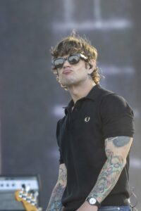 Male performer on stage wearing a black polo, large sunglasses, and a wristwatch, with colorful tattoo sleeves visible on both arms.