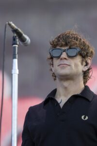 Musician wearing dark polo shirt and oversized sunglasses stands near a microphone on a stand outdoors.