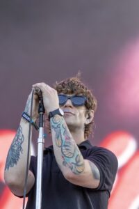 Male singer with tattoos on both forearms, wearing sunglasses, singing into a microphone on stage.