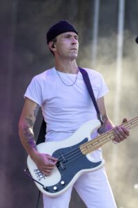 Male bassist in a black beanie and white t-shirt plays a white bass guitar on stage; visible tattoos on arms and a guitar strap across his chest.