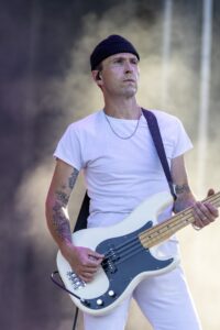 Male guitarist wearing a black beanie and white t-shirt plays a white bass guitar on stage outdoors, tattoos visible on his arms.