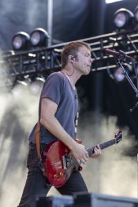 Male guitarist on stage playing a red electric guitar, wearing a gray t-shirt and in-ear monitors.