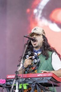 Male musician singing into a microphone, wearing a camo cap and green vest, holding tambourines on a pink-lit stage.