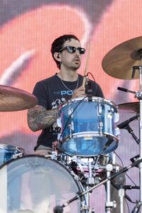Drummer wearing dark sunglasses playing a blue drum kit on stage with a red-lit background.