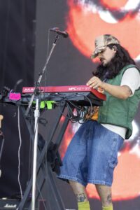 Male keyboardist performing on stage, wearing a camouflage cap and green vest, playing a red MPC Key37 keyboard with cables and mic stand nearby.