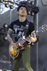 Male guitarist on stage wearing a black t‑shirt, playing a sunburst electric guitar with a decorative strap during a live performance.