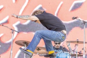Energetic drummer mid-air jumping while playing drums on stage, tattoos visible on forearm and microphone stands nearby.
